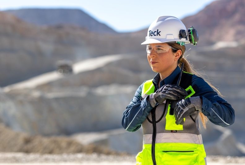 Engineer in safety gear stands at a mining site, hands clasped, with rocky terrain in background.